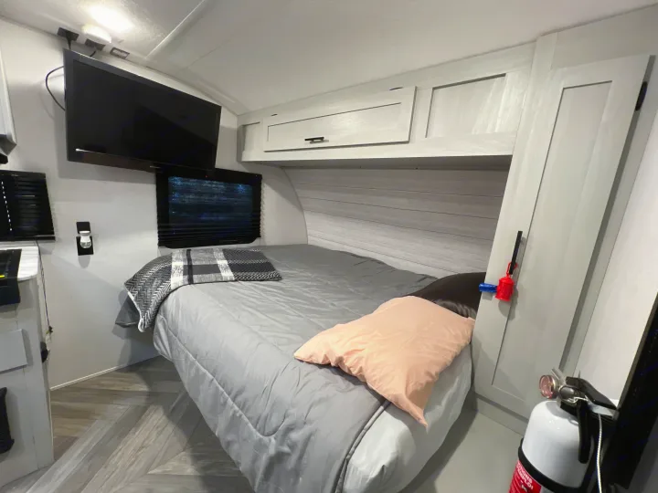 RV rental sleeping area featuring a queen-sized bed, wall-mounted TV, overhead cabinets, and a fire extinguisher.. Forest River Wildwood 2022