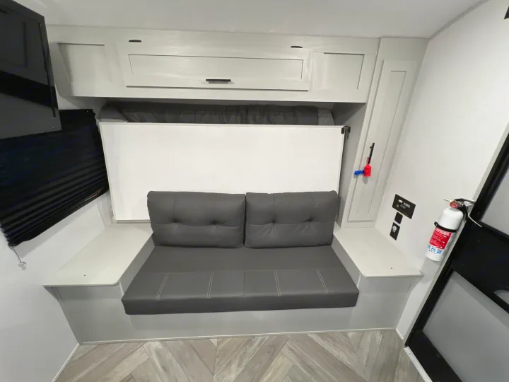 Inside an RV rental: a dark gray sofa with a folded murphy bed above, overhead cabinets, TV, and a fire extinguisher.. Forest River Wildwood 2022