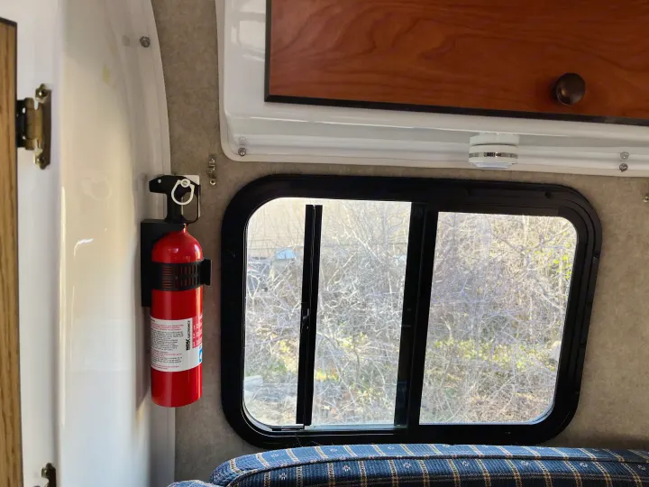 Essential RV rental safety: a fire extinguisher mounted beside a window with an outdoor view, cozy bed below.. Scamp Scamp Trailer 2004