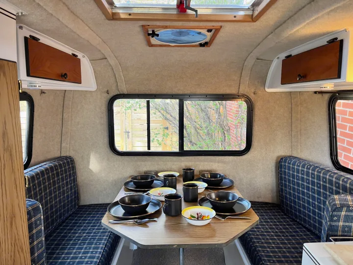 Cozy RV rental interior: dining area with a set table, blue plaid seats, windows, and overhead storage.. Scamp Scamp Trailer 2004