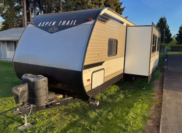 An Aspen Trail travel trailer (RV) with its slide-out extended, parked on sunny green grass. Perfect RV rental.. Aspen Trail Aspen Trail Trailer 2021