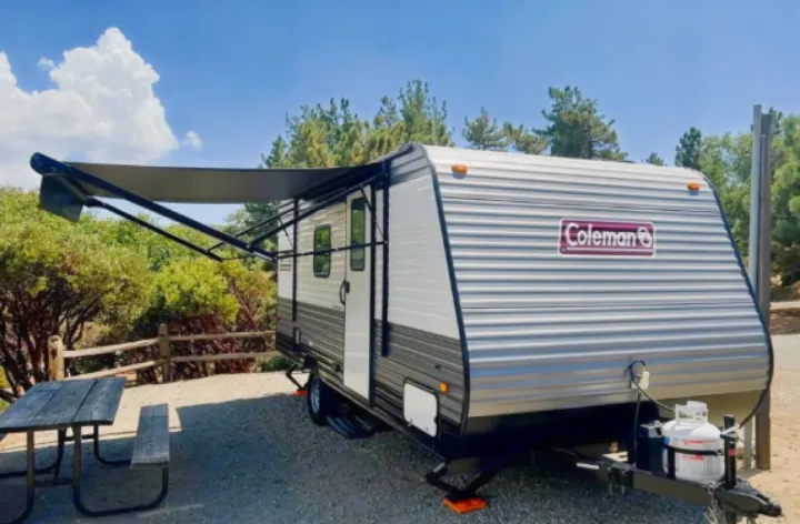 This RV rental features a travel trailer parked on a gravel campsite, with an extended awning and a picnic table nearby.. Coleman Lantern 17b 2024