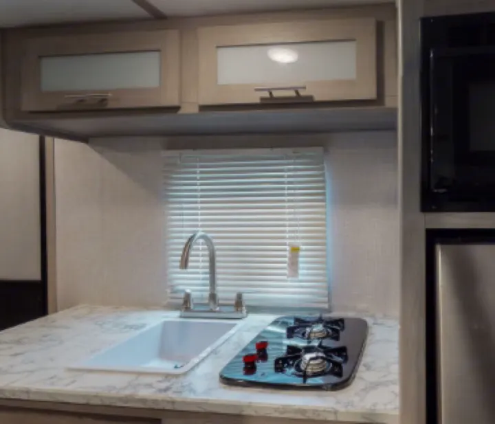 A bright RV rental kitchen: white countertop, steel sink, two-burner stove, and light wood cabinets with frosted glass.. Coleman Lantern 17b 2024