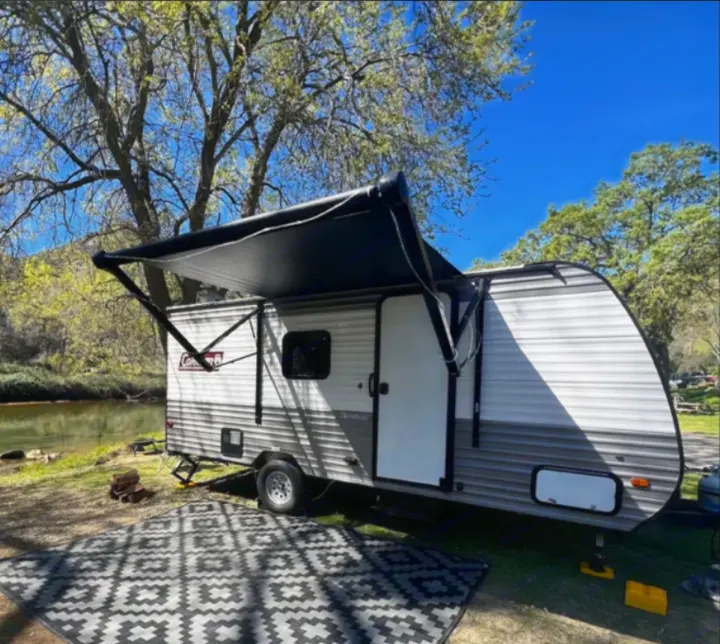 RV rental by a river with extended awning & patterned mat. Perfect for a peaceful camping trip surrounded by nature.. Coleman Lantern 17b 2024