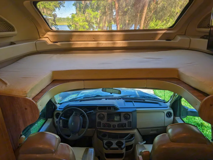 Interior of an rv rental: overhead bed, driver's dashboard, and a clear view of a lake and trees through the front window.. Jayco Greyhawk 2019