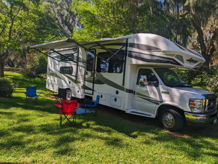 White Jayco rv rental with awning extended, parked on green grass with camping chairs & mat. Shaded by trees with Spanish moss.. Jayco Greyhawk 2019