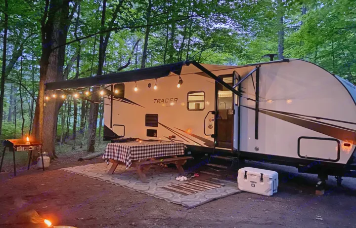 Evening RV rental campsite: cozy travel trailer under string lights, with picnic table, grill, cooler, and campfire.. Forest River Tracer LE 2020