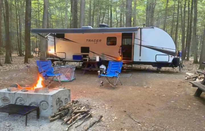 Cozy RV rental campsite featuring a lit campfire, blue chairs, and a white travel trailer with an extended awning in the woods.. Forest River Tracer LE 2020