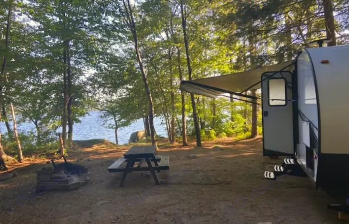 An rv rental is parked at a serene lakeside campsite with a picnic table and fire pit, surrounded by trees.. Forest River Tracer LE 2020