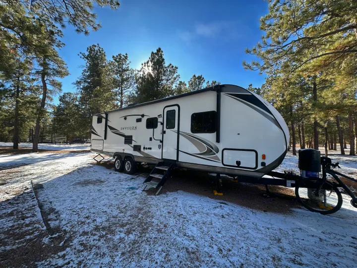 Enjoying a serene winter **rv rental** experience. A travel trailer is parked in a snowy pine forest campground under bright sun.. Gulf Stream Envision 2020
