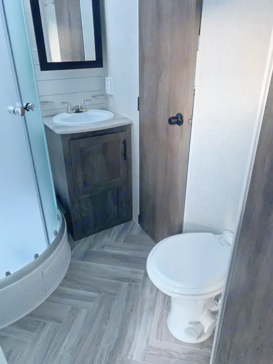 RV rental bathroom with a modern vanity, mirror, toilet, and curved shower, all on stylish wood-look flooring.. Forest River Stealth 2021