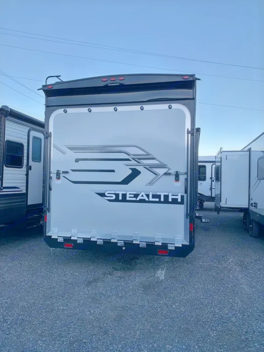A "Stealth" brand RV ready for rental. The back shows a large grey ramp door, parked on a gravel lot among other RVs.. Forest River Stealth 2021