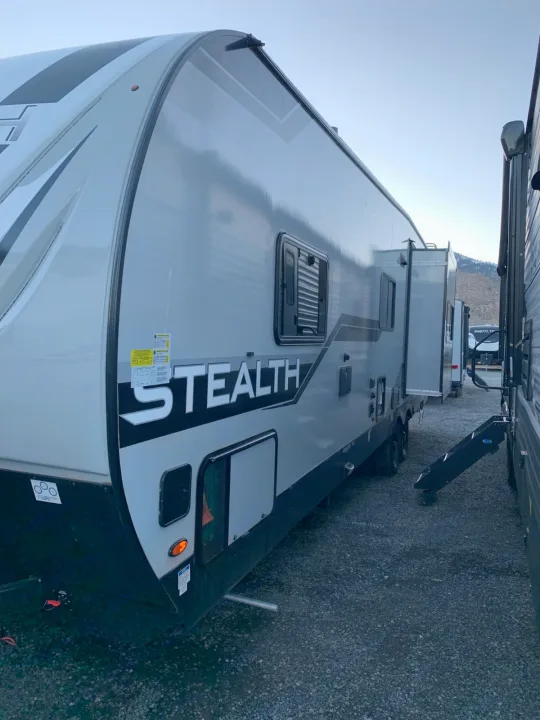 A grey Stealth RV rental, a travel trailer with an extended slide-out, parked on gravel.. Forest River Stealth 2021
