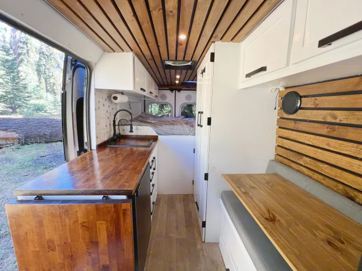 View inside a modern RV rental camper van showing a bright kitchen with wood counters, a dining area, and a rear bed. Trees are visible outside.. Dodge Sprinter Van 2019