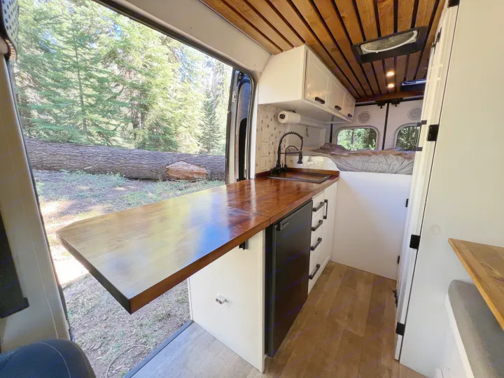 Spacious RV rental interior with a fully equipped kitchen, cozy bed, and an expansive forest view through the open side door.. Dodge Sprinter Van 2019