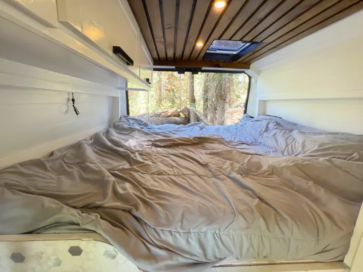 Cozy RV rental bed with white shiplap walls, wooden ceiling, and a scenic forest view from the back window.. Dodge Sprinter Van 2019