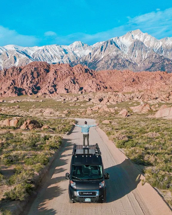 A person on a black RV rental faces majestic snow-capped mountains & red rock formations on a dirt road.. Dodge Sprinter Van 2019