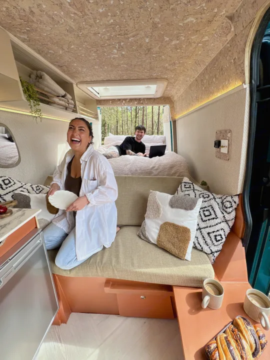 Enjoying a cozy RV rental: a woman laughs by the kitchen, and a man works on a laptop from the bed, with forest views.. Mercedes-Benz Sprinter 2016