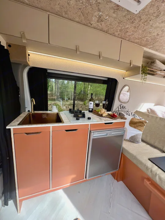 Stylish RV rental interior: a modern orange kitchen with sink, stovetop, and fridge, next to a window showing a forest.. Mercedes-Benz Sprinter 2016