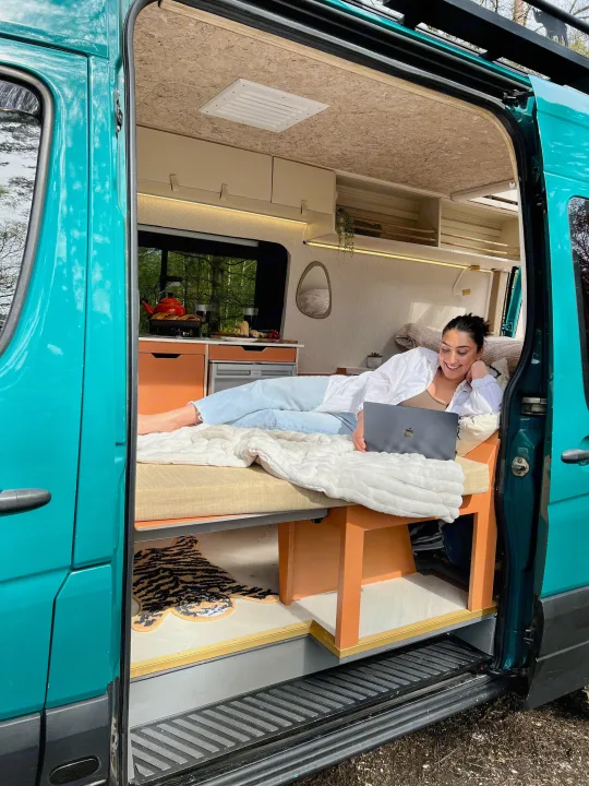 A woman works on her laptop inside a teal **rv rental**, lounging on a cozy bed with a full kitchen visible.. Mercedes-Benz Sprinter 2016
