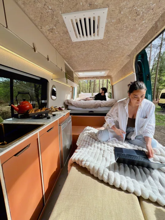 Bright RV rental: a woman on a cozy blanket with laptop, a man on the bed, and a modern kitchen. Perfect for van life.. Mercedes-Benz Sprinter 2016