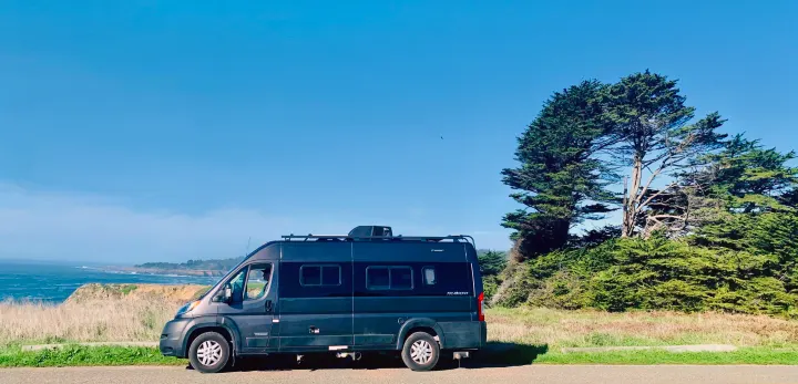 Dark grey Winnebago Travato RV rental parked on a coastal road overlooking ocean cliffs and green trees under a clear sky.. Winnebago Travato 2022