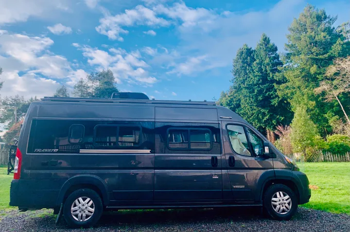 Dark gray Winnebago Travato RV rental, a modern camper van, parked on gravel next to green trees under a blue sky.. Winnebago Travato 2022