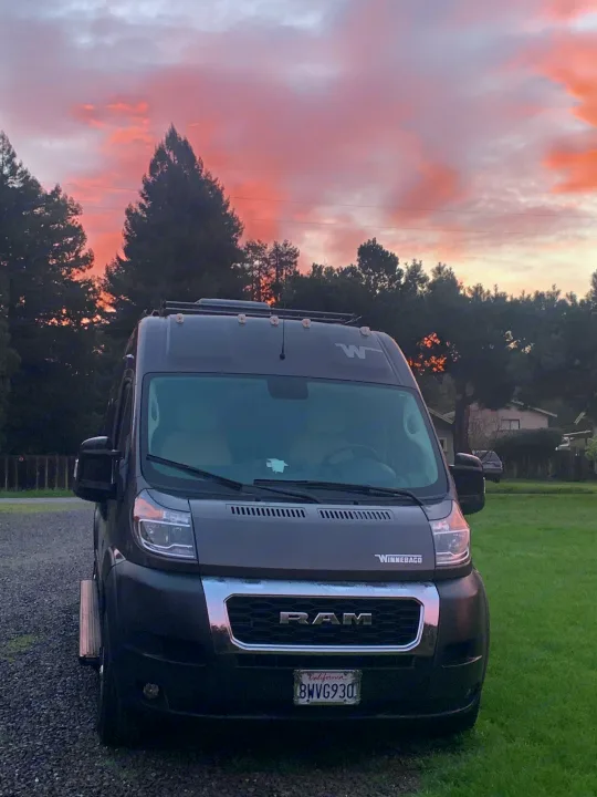 Dark gray Winnebago RV rental van parked at sunset with vibrant pink and orange clouds overhead. Gravel and grass visible.. Winnebago Travato 2022