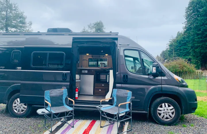 Experience comfort with this dark grey camper van RV rental. Open door shows interior kitchen, two chairs on rug outside.. Winnebago Travato 2022