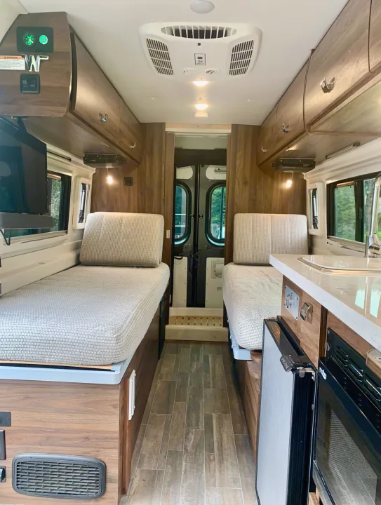 Bright RV rental interior with twin beds, a compact kitchen (sink, fridge, oven), and overhead storage.. Winnebago Travato 2022