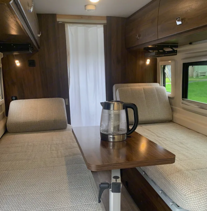 RV rental interior features light fabric bench seating/beds, a central adjustable table with a kettle, and overhead cabinets.. Winnebago Travato 2022