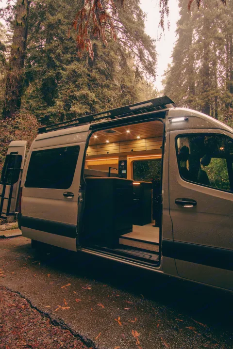 Modern camper van, perfect for an rv rental adventure, parked in a lush forest with its illuminated interior visible.. Mercedes-Benz Sprinter 2023
