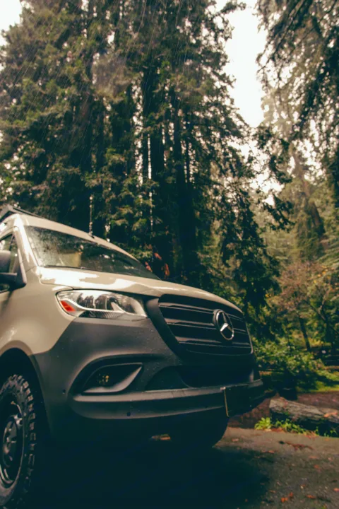 Experience nature with this RV rental. A modern van in a wet, dense forest, rain streaking the tall trees.. Mercedes-Benz Sprinter 2023