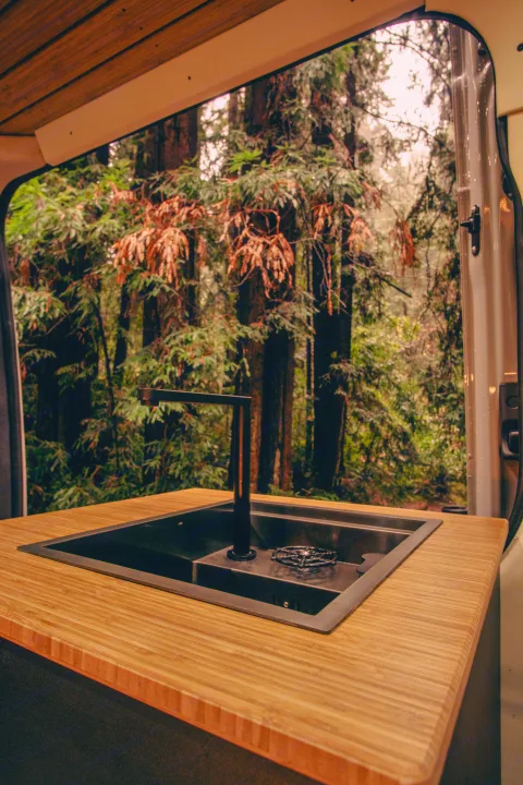 RV rental camper van interior: sleek black sink, wood counter, and a beautiful forest view from the open door.. Mercedes-Benz Sprinter 2023