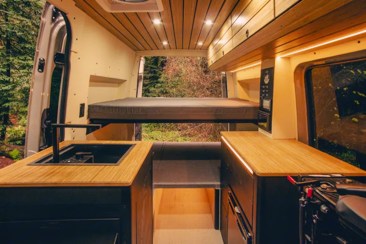Inside a custom RV rental, this spacious camper van features a raised bed, kitchen, and forest views from the open rear.. Mercedes-Benz Sprinter 2023