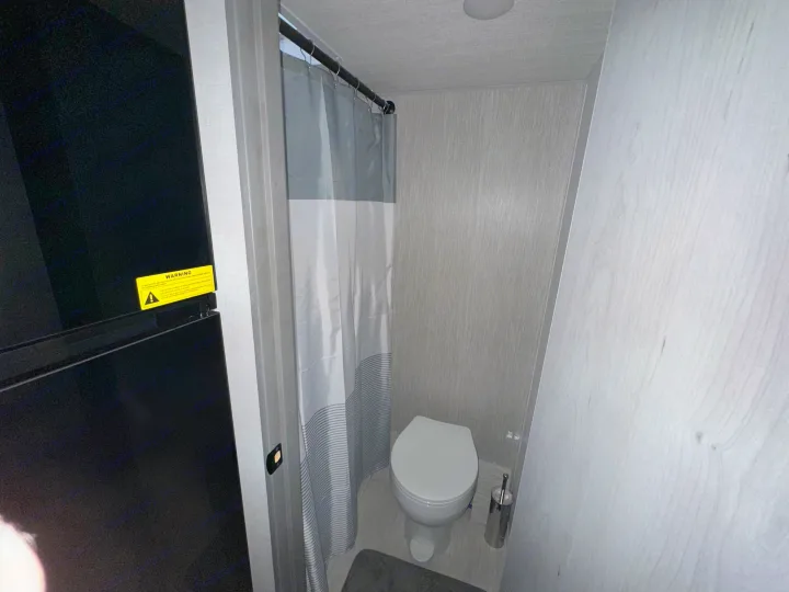 Inside an RV rental: a compact bathroom with a white toilet, gray shower curtain, and a large black appliance on the left.. Forest River Ozark 2024
