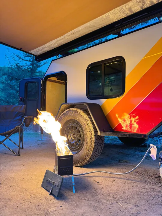 Cozy evening camping with an rv rental. Portable propane campfire lights up the striped teardrop camper under its awning.. Custom Retro 2023