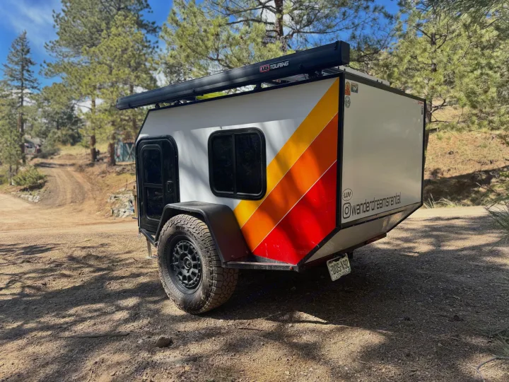 This unique off-road camper RV rental, featuring colorful stripes & an awning, is parked on a dirt trail amidst pine trees.. Custom Retro 2023