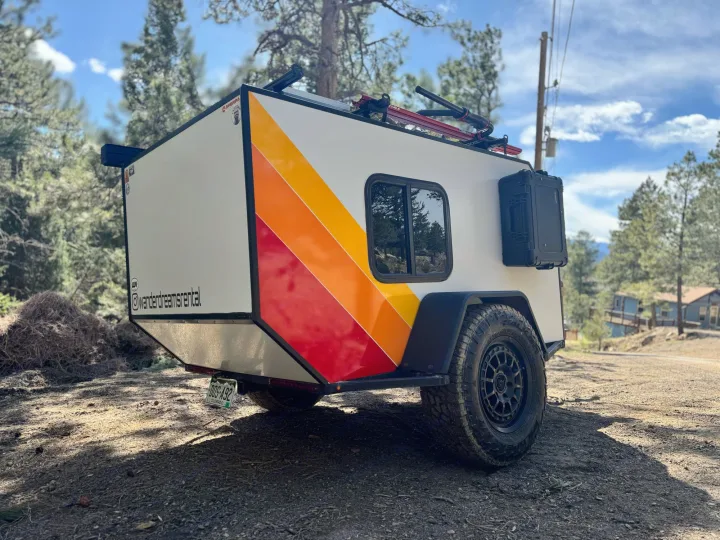 White teardrop RV rental by Wander Dreams, with vibrant stripes & off-road tires, on a dirt road for adventure.. Custom Retro 2023