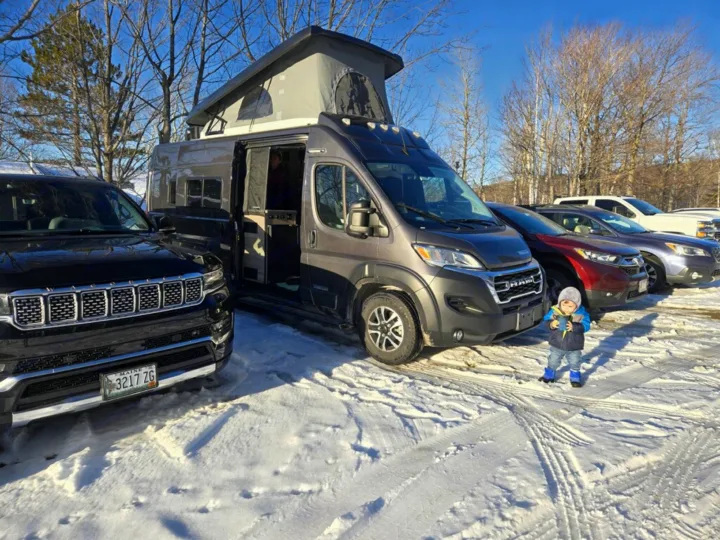 Pop-top RV rental van in a snowy parking lot with other cars and a child. Ready for winter travel adventures!. Winnebago Solis 2023