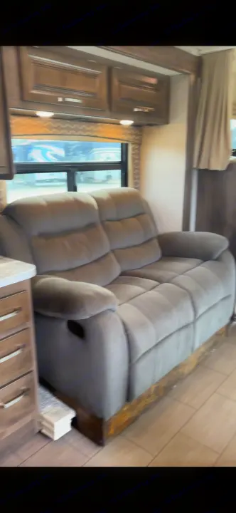 Cozy RV rental interior featuring a plush gray recliner sofa, wooden overhead cabinets, and a large window.. Jayco Greyhawk 2019
