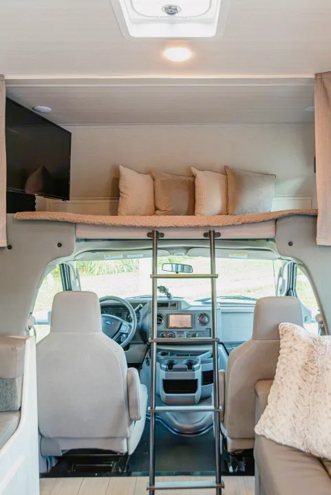 Bright rv rental interior looking into the Ford cab, with an inviting overhead bunk bed, soft pillows, and access ladder.. East to West Entrada 2024