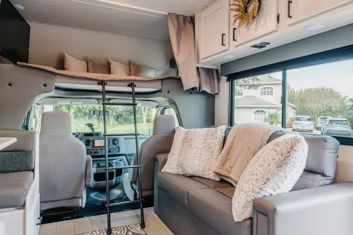 Cozy RV rental interior featuring an over-cab bunk bed, plush sofa, dinette, and cockpit view.. East to West Entrada 2024