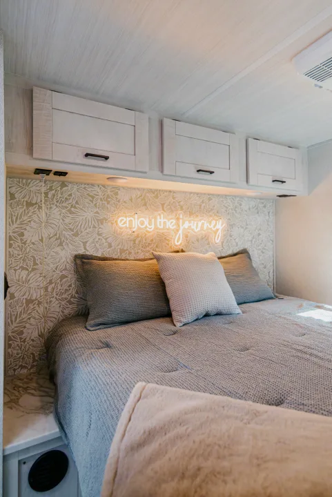 Cozy RV rental bedroom with "enjoy the journey" neon sign, waffle-knit bedding, and overhead storage for comfort on the road.. East to West Entrada 2024