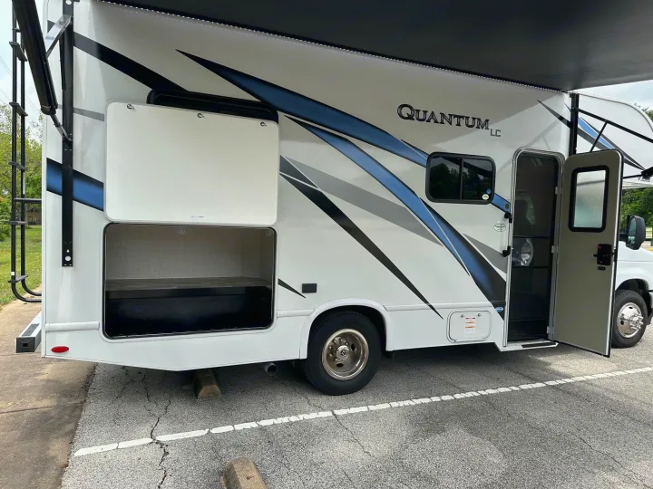White rv rental motorhome with blue/black graphics, showing open storage & entry doors, parked on asphalt.. Thor Quantum LC 2024