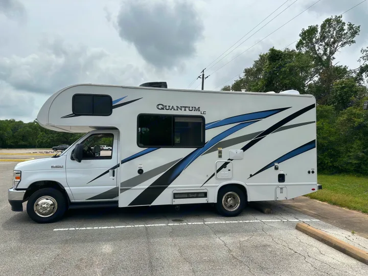 A white Quantum LC rv rental, a Class C motorhome, parked on asphalt under cloudy skies, with trees behind. Blue, black, gray accents.. Thor Quantum LC 2024