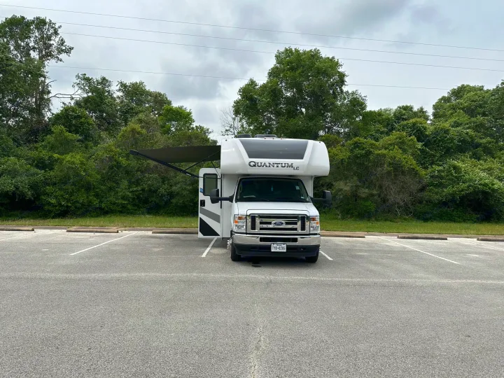 White Ford Quantum LC RV rental motorhome parked in a lot surrounded by green trees under a cloudy sky.. Thor Quantum LC 2024