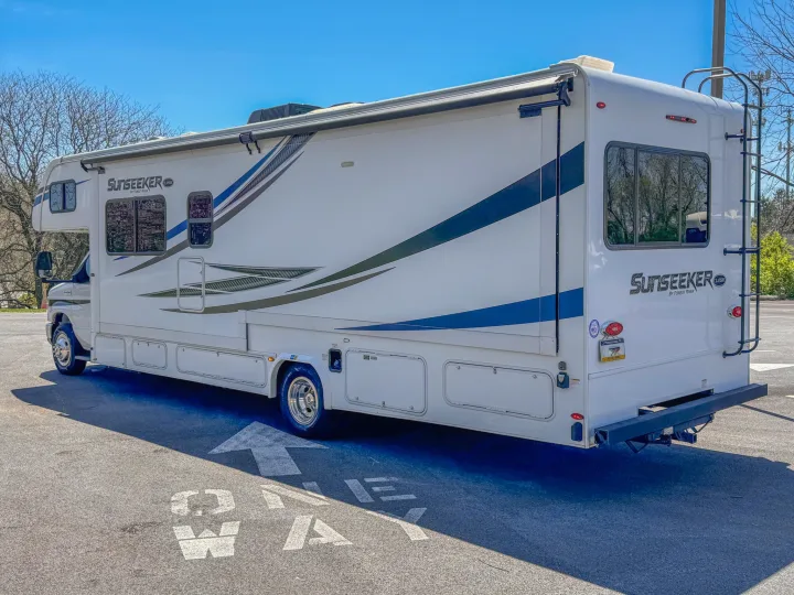 Large white Sunseeker RV rental with blue stripes, parked on asphalt, showing its side awning and rear ladder.. Forest River Sunseeker 2021