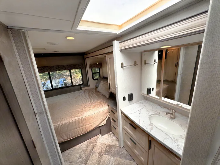 RV rental interior: A bright bedroom with a bed & windows, plus a skylight, connects to a vanity with marble sink & mirror.. Forest River Sunseeker 2021