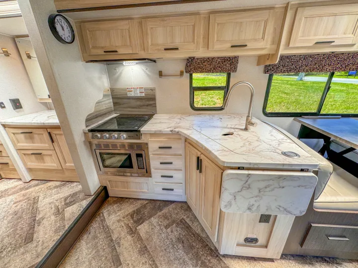 Modern RV rental kitchen interior, featuring light wood cabinets, stone-look countertops, stove, microwave, and sink.. Forest River Sunseeker 2021